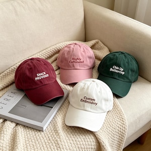 Può includere: Quattro cappellini da baseball in bordeaux, rosa, verde scuro e bianco. Il cappellino bordeaux presenta il testo SNACK PROVIDER, quello rosa dice Highly Caffeinated, quello verde scuro recita Chin Up Buttercup e quello bianco Custom Embroidery.