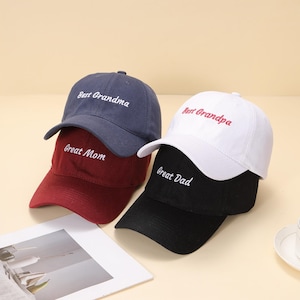 Puede incluir: Tres gorras de béisbol en azul marino, burdeos, blanco y negro. Cada gorra tiene texto bordado: "Best Grandma", "Great Mom", "Best Grandpa" y "Great Dad". Las gorras están apiladas, con las viseras hacia adelante.