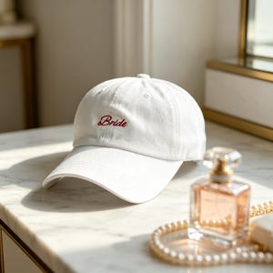 Puede incluir: Gorra de béisbol blanca con la palabra "Bride" bordada en rojo. La gorra tiene una visera curva y un botón en la parte superior. Se encuentra sobre una superficie de mármol.