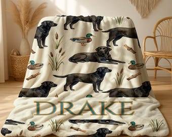 May include: Cream-colored throw blanket featuring a repeating pattern of black Labrador Retrievers, ducks, and reeds. The word "DRAKE" is displayed in a teal and gold font across the blanket. The blanket is draped over a chair.