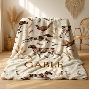 Puede incluir: Manta crema con un estampado repetido de perros marrones y blancos, faisanes y espigas de trigo. La palabra "GABLE" est&aacute; impresa en marr&oacute;n con una fuente estilizada. La manta est&aacute; hecha de un material suave y afelpado.