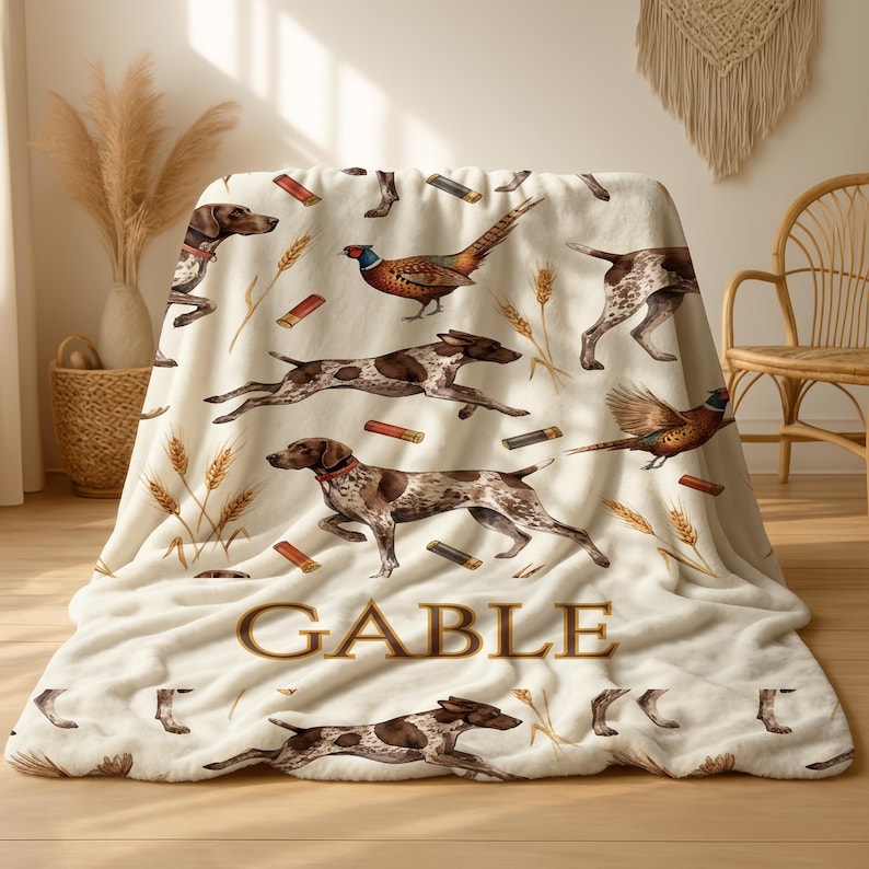 Puede incluir: Manta crema con un estampado repetido de perros marrones y blancos, faisanes y espigas de trigo. La palabra "GABLE" est&aacute; impresa en marr&oacute;n con una fuente estilizada. La manta est&aacute; hecha de un material suave y afelpado.