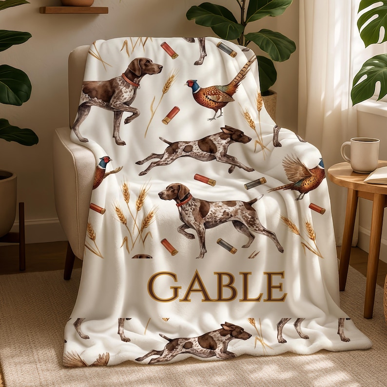 Puede incluir: Una manta blanca con un patr&oacute;n repetido de perros marrones y blancos, faisanes, espigas de trigo y cartuchos de escopeta. El nombre "GABLE" est&aacute; impreso en letras doradas. La manta est&aacute; sobre un sill&oacute;n beige.
