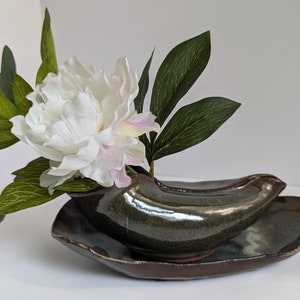 May include: A white peony flower with green leaves arranged in a dark green ceramic vase. The vase is shaped like a banana and sits on a dark brown ceramic dish.