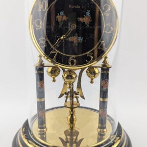 May include: A gold-toned Kundo pendulum clock with a black face and floral accents. The clock is encased in a glass dome.
