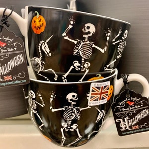 May include: Three black ceramic mugs with white skeleton designs and a pumpkin. The mugs have white rims and handles. The mugs have tags that say "Designed in England" and "Halloween" with a red bird logo.