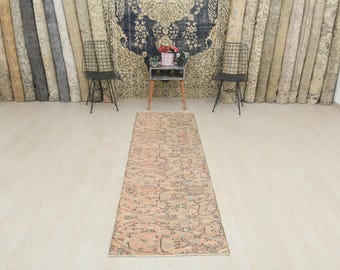 Puede incluir: Una alfombra rectangular de estilo vintage con un diseño floral descolorido en tonos melocotón, beige y gris. La alfombra es larga y estrecha, adecuada para un pasillo o entrada. El fondo incluye alfombras enrolladas y elementos decorativos.