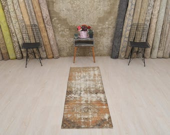 May include: A vintage style rug with a faded, distressed look in shades of beige, brown, and cream. The rug is laid out on a light wood floor in front of a wall with a row of decorative fabric rolls.