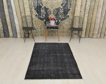Puede incluir: Una alfombra rectangular negra con un patrón descolorido y desgastado está centrada en un suelo de madera clara. La alfombra es el punto focal, con una pequeña mesa y dos sillas en el fondo. Alfombras enrolladas alinean la pared.