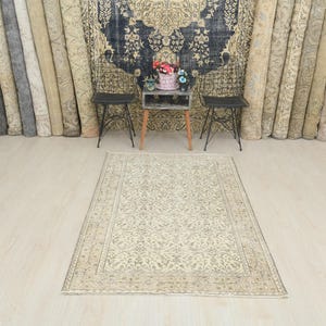 Puede incluir: Una alfombra rectangular beige con un intrincado diseño floral. La alfombra está centrada en un suelo de madera clara. Alfombras enrolladas bordean la pared trasera, con una pequeña mesa y dos sillas en el fondo.