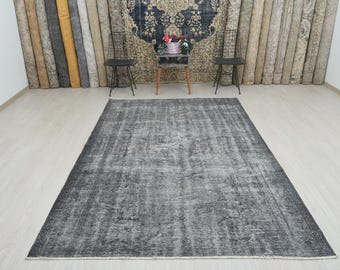 May include: A large, rectangular, distressed gray rug with a faded, vintage appearance. The rug features a central design with lighter gray accents and a darker gray border. The rug is placed on a light-colored wooden floor.