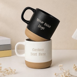 May include: A black ceramic mug with the text 'your own Logo' rests on top of a white speckled ceramic mug with the text 'Custom Text Here'. Both mugs feature a natural tan unglazed base and a curved handle, showcasing a two-tone design.