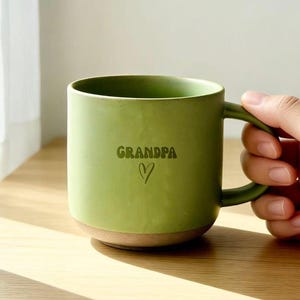 Peut inclure: Une tasse en céramique vert clair mat avec une anse arrondie. L'avant de la tasse présente le mot "GRANDPA" imprimé dans une police rétro vert foncé, positionné au-dessus d'un contour de cœur simple dessiné à la main en vert foncé.