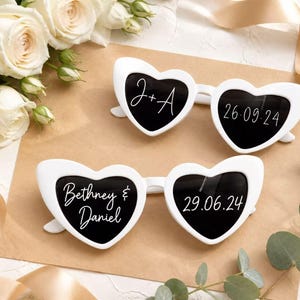 May include: Two pairs of white heart-shaped sunglasses with black lenses. The top pair features the text J+A and 26.09.24. The bottom pair features the text Bethney & Daniel and 29.06.24 printed in white script on the lenses.