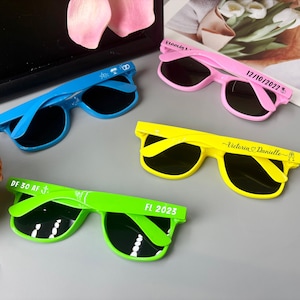 May include: Four pairs of sunglasses in blue, pink, yellow, and green. Each pair has dark lenses and text on the arms. The pink pair says "Kennedy S" and "12/10/2022". The yellow pair says "Victoria Danielle". The green pair says "DF 30 AF" and "FL 2023".