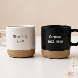 Può includere: Due tazze in ceramica con finitura maculata, una bianca e una nera, entrambe con base color terra. La tazza bianca riporta la scritta 'Your Own Logo' e la tazza nera riporta la scritta 'Custom Text Here' in un semplice carattere scuro senza grazie.