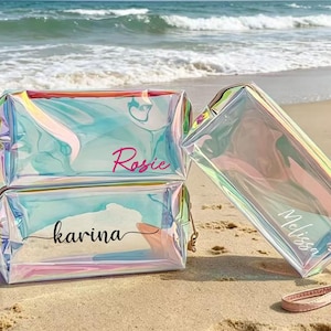 May include: Iridescent cosmetic bags with names 'Rosie', 'Karina', and 'Melissa' printed on them. The bags are rectangular with a zipper closure and a wrist strap. The bags are on a sandy beach with the ocean in the background.