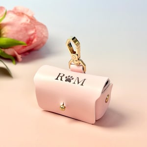 May include: A pale pink leather pet waste bag dispenser with a gold-coloured clasp. The dispenser has a paw print and the initials "R M" printed on it. A pink rose is in the background.