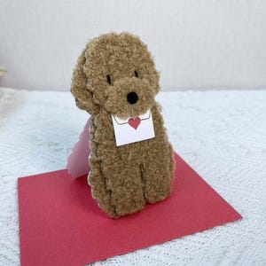 May include: A brown teddy bear with a fluffy texture is holding a white envelope with a red heart on it. The bear is standing on a red card.