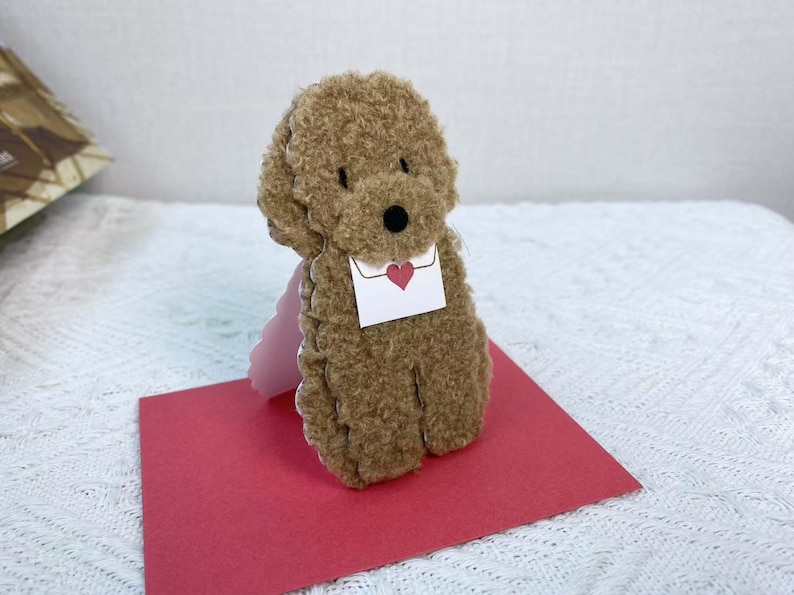 May include: A brown teddy bear with a fluffy texture is holding a white envelope with a red heart on it. The bear is standing on a red card.