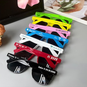 May include: A collection of sunglasses in various colors, including green, yellow, pink, blue, white, red, and black. Each pair has dark lenses and white text on the frames, with names and dates. The sunglasses are arranged in a diagonal row.