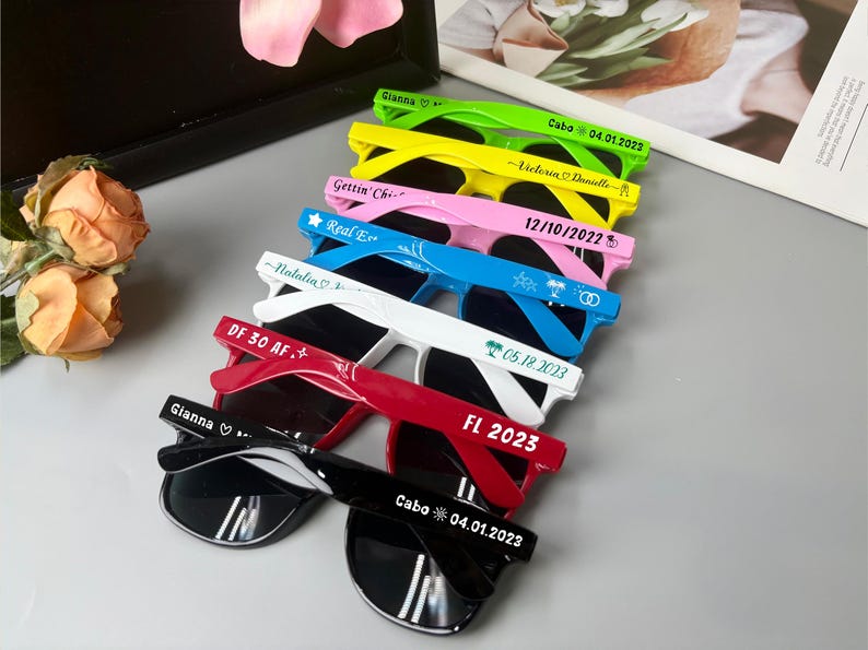 May include: A collection of sunglasses in various colors, including green, yellow, pink, blue, white, red, and black. Each pair has dark lenses and white text on the frames, with names and dates. The sunglasses are arranged in a diagonal row.