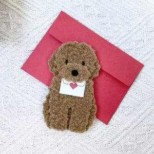 May include: A brown plush toy poodle holding a white envelope with a red heart on it. The poodle is sitting on a red envelope.
