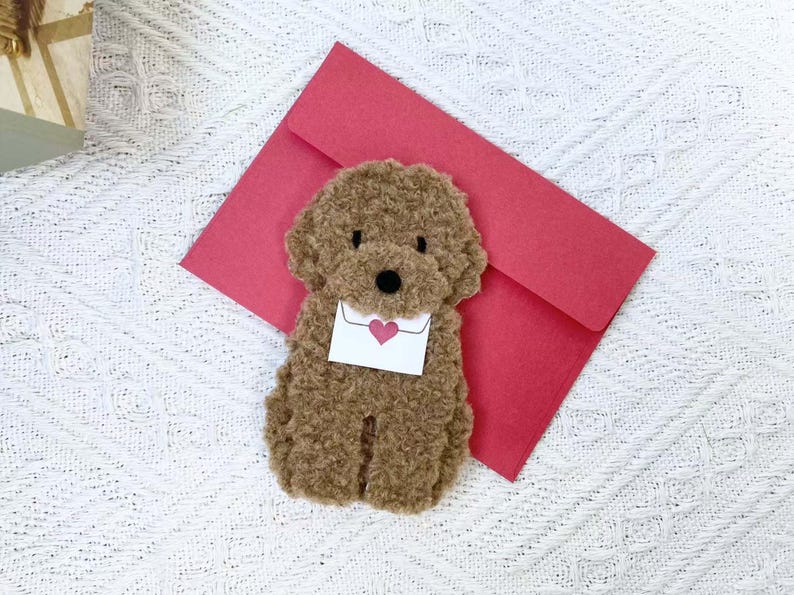 May include: A brown plush toy poodle holding a white envelope with a red heart on it. The poodle is sitting on a red envelope.