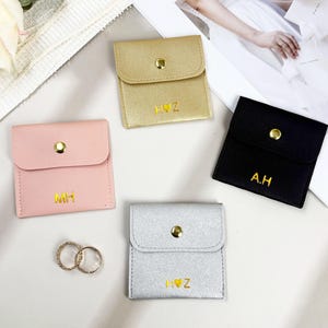May include: Four small, square pouches in pink, gold, black, and silver. Each pouch has a flap closure with a gold snap and gold lettering. Two rings are next to the pink pouch. The pouches are for jewelry.