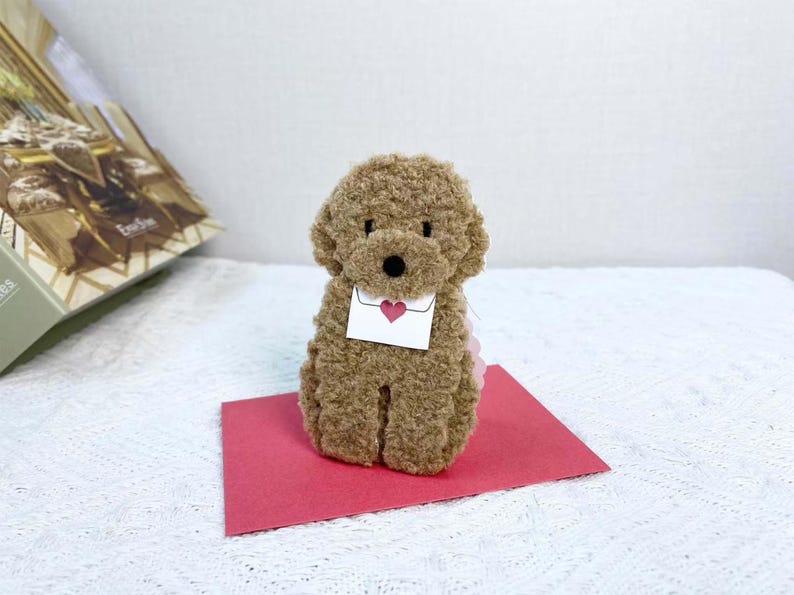May include: A brown plush toy poodle holding a white envelope with a red heart on it. The poodle is sitting on a red card.