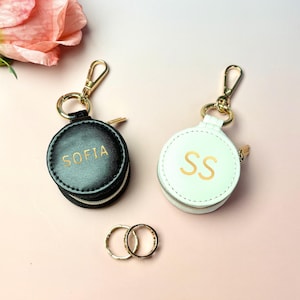 May include: Two round, zippered keychains, one black with "SOFIA" in gold lettering, the other white with "SS". Both have gold-colored hardware. Two gold rings are below them, and a pink rose is in the background.