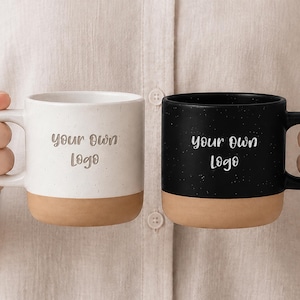 May include: Two ceramic mugs, one white and one black, both featuring a speckled finish and a natural terracotta-colored base. Each mug has the text 'your own logo' printed in a simple font on the front, with a handle on the side of each.