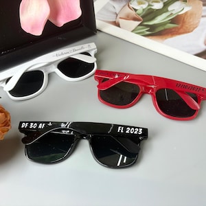 May include: Three pairs of sunglasses in white, red, and black. The white sunglasses have "Victoria Danielle" printed on the arm. The red sunglasses have "Gettin' Hitched" and "12/10/2022" printed on the arm. The black sunglasses have "DF 30 AF" and "FL 2023" printed on the arm.