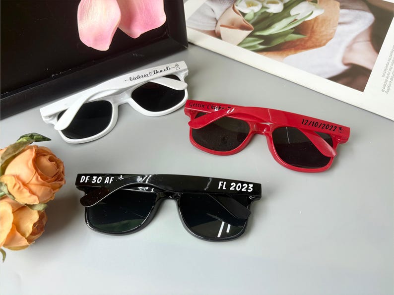 May include: Three pairs of sunglasses in white, red, and black. The white sunglasses have "Victoria Danielle" printed on the arm. The red sunglasses have "Gettin' Hitched" and "12/10/2022" printed on the arm. The black sunglasses have "DF 30 AF" and "FL 2023" printed on the arm.