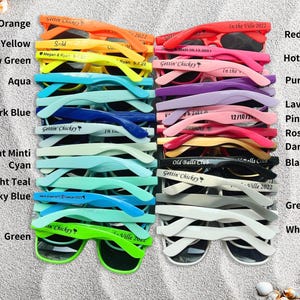 May include: A collection of sunglasses in various colors, including orange, yellow, aqua, dark blue, and green. Each pair has a unique inscription. The sunglasses are arranged on a sandy surface.