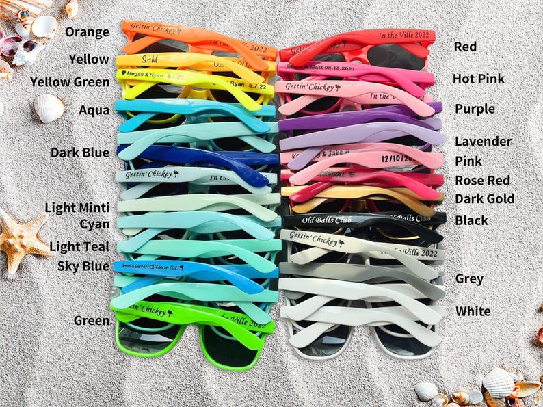 May include: A collection of sunglasses in various colors, including orange, yellow, aqua, dark blue, and green. Each pair has a unique inscription. The sunglasses are arranged on a sandy surface.