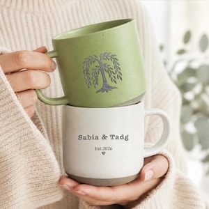 Peut inclure: Une pile de deux tasses en céramique. La tasse supérieure est vert sauge avec une illustration d'arbre saule gris foncé. La tasse inférieure est blanc cassé avec le texte gris foncé "Sabia & Tadg", "Est.2026" et un petit cœur imprimé.