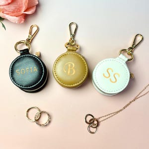 May include: Three round coin purses with gold-coloured keyrings, in black, gold, and white. The black purse has "SOFIA" in gold lettering, the gold purse has a gold "B", and the white purse has "SS". Gold jewellery, including rings and a necklace, is also visible.