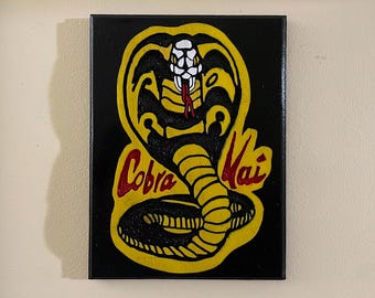 Puede incluir: Una pintura sobre lienzo negro con una cobra amarilla con lengua roja y las palabras "Cobra Kai" en letras rojas.