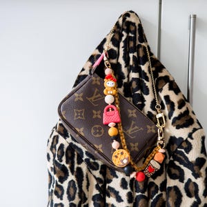 May include: A brown leather handbag with a gold chain strap and a decorative charm. The charm features a variety of colorful beads and a small pink purse. The handbag is hanging on a leopard print coat.