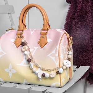 Peut inclure: Sac à main dégradé rose et jaune avec un motif floral blanc. Le sac a des poignées et des accents en cuir beige. Un charm décoratif en perles avec des perles en bois, des fleurs et un nuage est suspendu à la poignée.