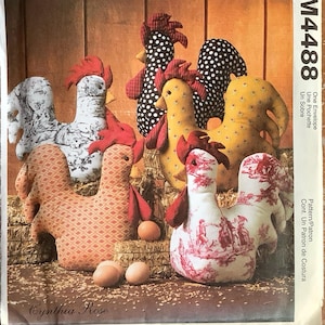 May include: McCall's Crafts pattern M4488 featuring various fabric rooster designs. The roosters are made of patterned fabrics in red, white, black, and yellow. The pattern includes instructions for creating stuffed rooster decorations.