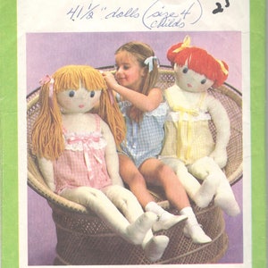 May include: Simplicity pattern 8967 features instructions for making 4.5-inch dolls. The image shows a child with two dolls in a wicker chair. The dolls have yarn hair and are wearing dresses. The pattern is marked "One Size."