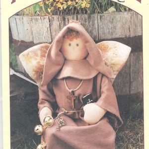 May include: A brown fabric doll dressed as a monk with a brown hooded robe, a rope belt, a cross necklace, and a book. The doll has a wing on its back and is holding a book in its hand. The doll is sitting on a bed of green grass. The doll is 22 inches tall. The image is from a pattern book titled "Twice As Nice Designs, LTD: A Wing & A Prayer No. 218"