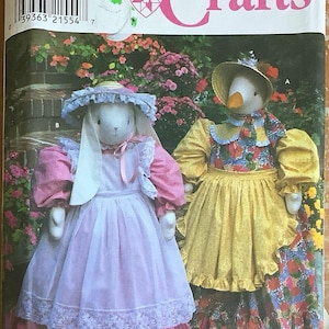 May include: Simplicity Crafts pattern featuring two stuffed animal dolls: a bunny in a pink dress and a goose in a yellow dress. The pattern is labeled "8128" and "ONE SIZE". The background shows a garden with pink flowers. The pattern is by Faith Van Zanten.