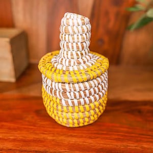 May include: A small, round, woven basket with a lid. The basket is made from natural materials and is decorated with yellow and white stripes.