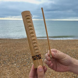 Könnte beinhalten: Ein handgefertigter Regenstab aus Bambus mit einem geschnitzten Muster und einem dekorativen Band. Ein Holzstab wird daneben gehalten. Im Hintergrund ein Strand, das Meer und der Himmel.