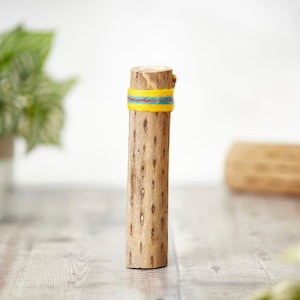 May include: A cylindrical wooden object with a textured surface. A band of yellow, blue, and red yarn is wrapped around the top. The object is standing upright on a light-colored wooden surface.