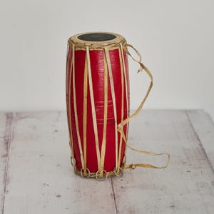Op de afbeelding: Een rode handtrommel met een houten lichaam en een weefsel van natuurlijke vezels. De trommel heeft een leren kop en is vastgebonden met een touw van natuurlijke vezels.