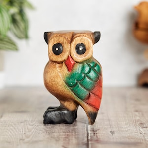 Può includere: Una statuetta di gufo in legno intagliata a mano, dipinta di verde, rosso e marrone. Il gufo ha grandi occhi neri e un becco.